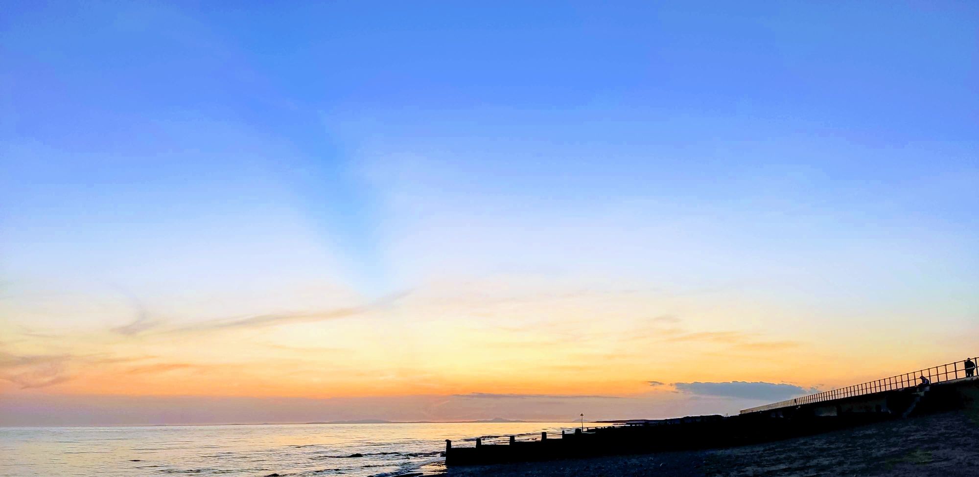 A wide evening sky fading from blue to gold over the sea, with the promenade wall and groynes in silhouette
