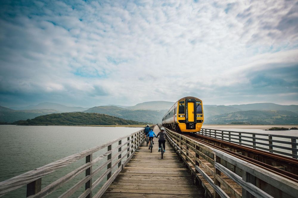 A train crossing a wooden estuary bridge, with cyclists on the walkway beside it