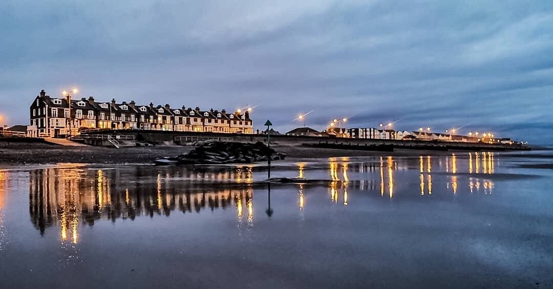 The sea front terrace at dusk, its lit windows and street lamps reflected in the wet sand of the beach